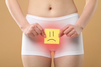 Image of Woman holding sticky note with drawn sad face and suffering from cystitis symptoms on beige background, closeup