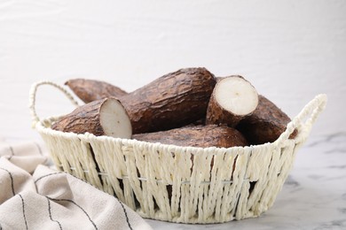 Photo of Whole and cut cassava roots in wicker basket on white marble table
