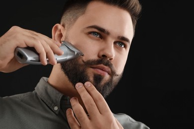 Handsome young man trimming beard on black background