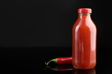 Photo of Spicy chili sauce in bottle and pepper against dark background, space for text