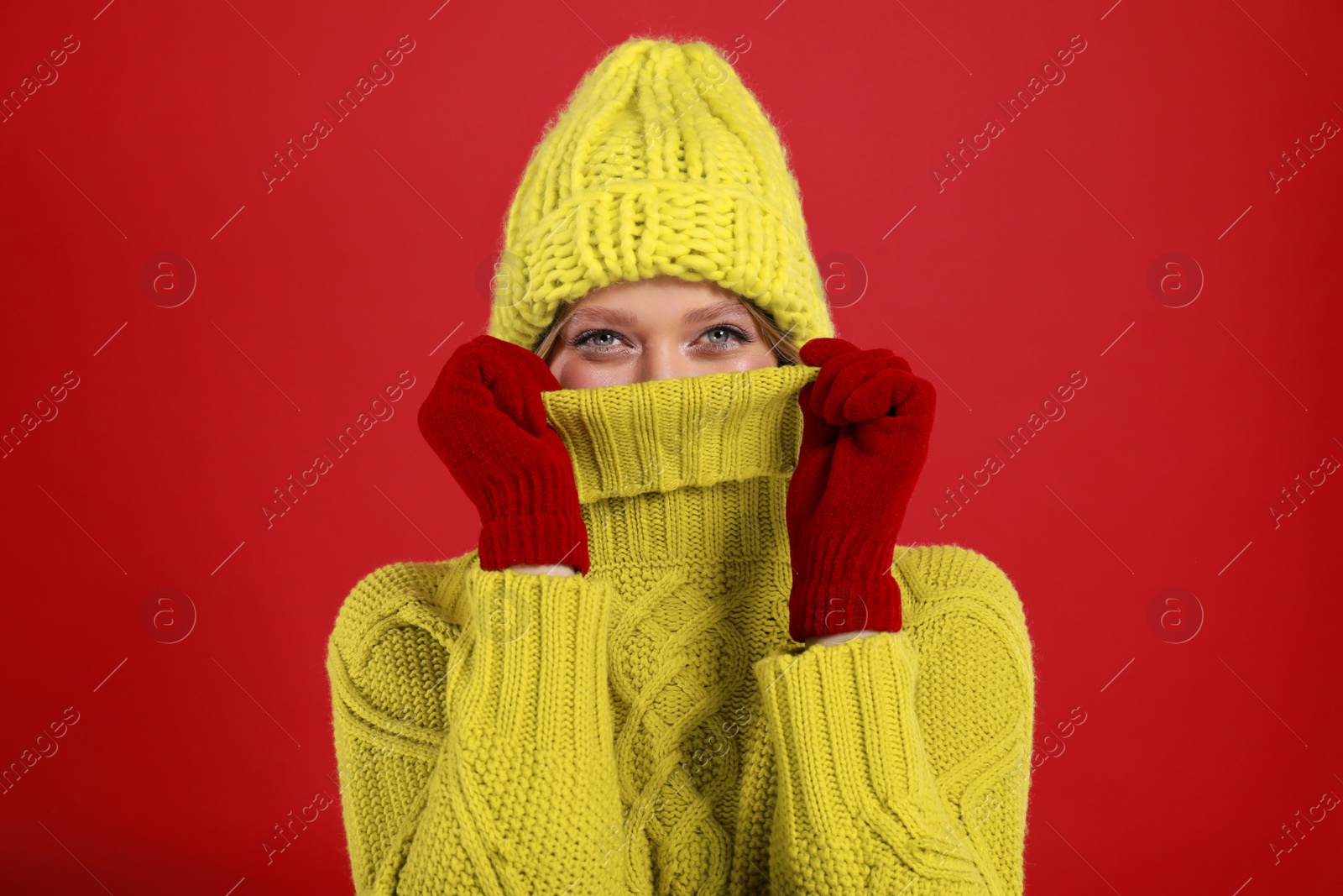 Photo of Funny young woman in warm sweater, gloves and hat on red background. Winter season
