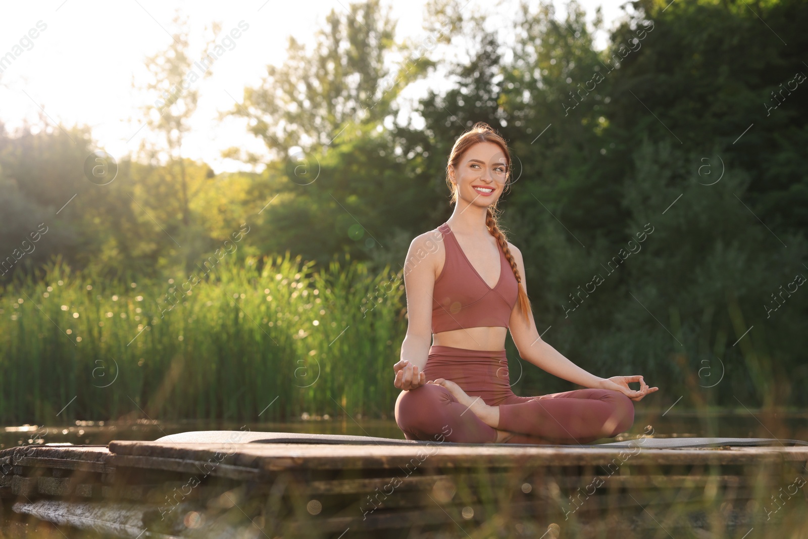 Photo of Beautiful happy woman practicing Padmasana on yoga mat on wooden pier near pond, space for text. Lotus pose