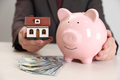 Man holding house model, piggy bank and dollar banknotes at wooden table, closeup. Saving money concept