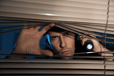 Photo of Male security guard with flashlight looking through window blinds in darkness