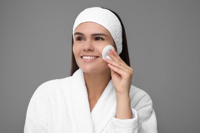 Young woman cleaning her face with cotton pad on grey background