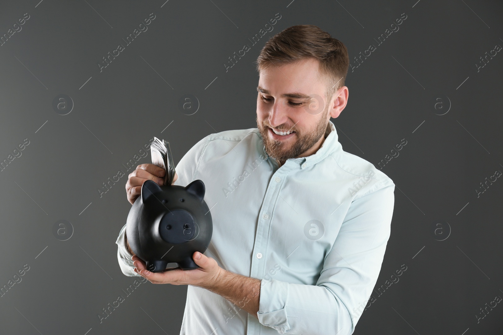 Photo of Happy young man putting money into piggy bank on grey background
