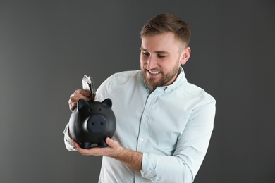 Happy young man putting money into piggy bank on grey background