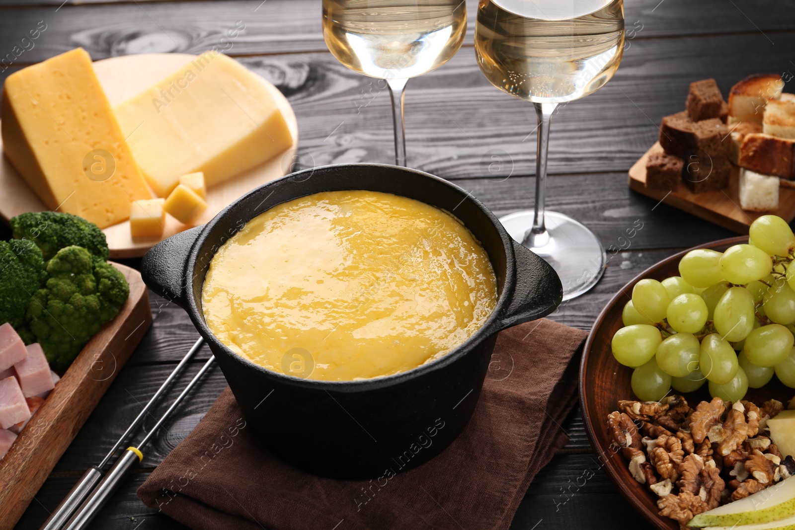 Photo of Fondue pot with melted cheese, glasses of wine and different products on black wooden table