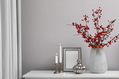 Photo of Hawthorn branches with red berries in vase, candles and frame on table indoors, space for text