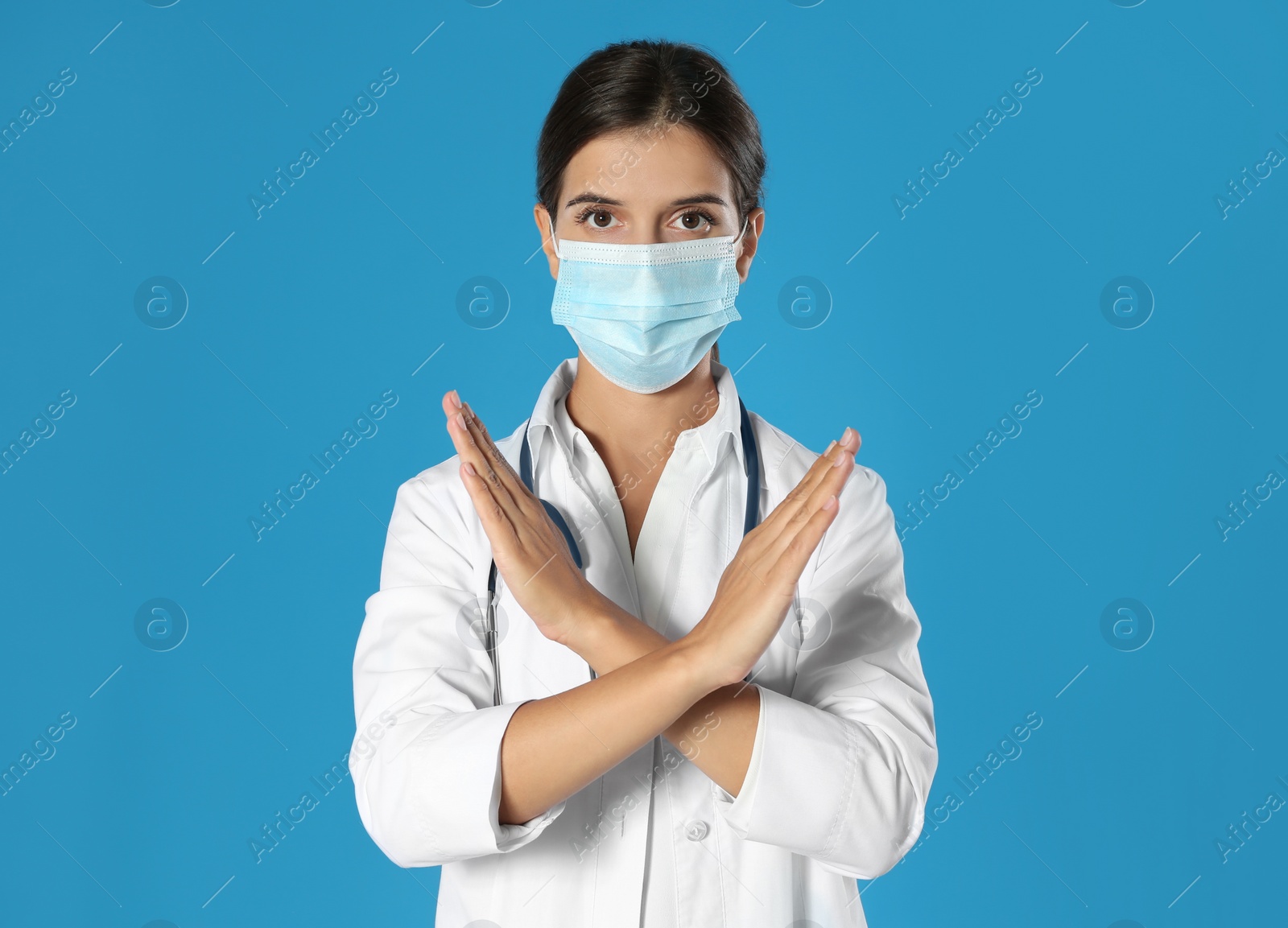 Photo of Doctor in protective mask showing stop gesture on blue background. Prevent spreading of coronavirus