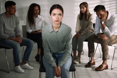 Photo of Unhappy young woman and group of people behind her back indoors. Therapy session