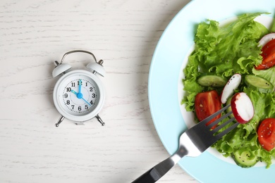 Photo of Alarm clock and plate with salad on white wooden 
table, flat lay. Meal timing concept