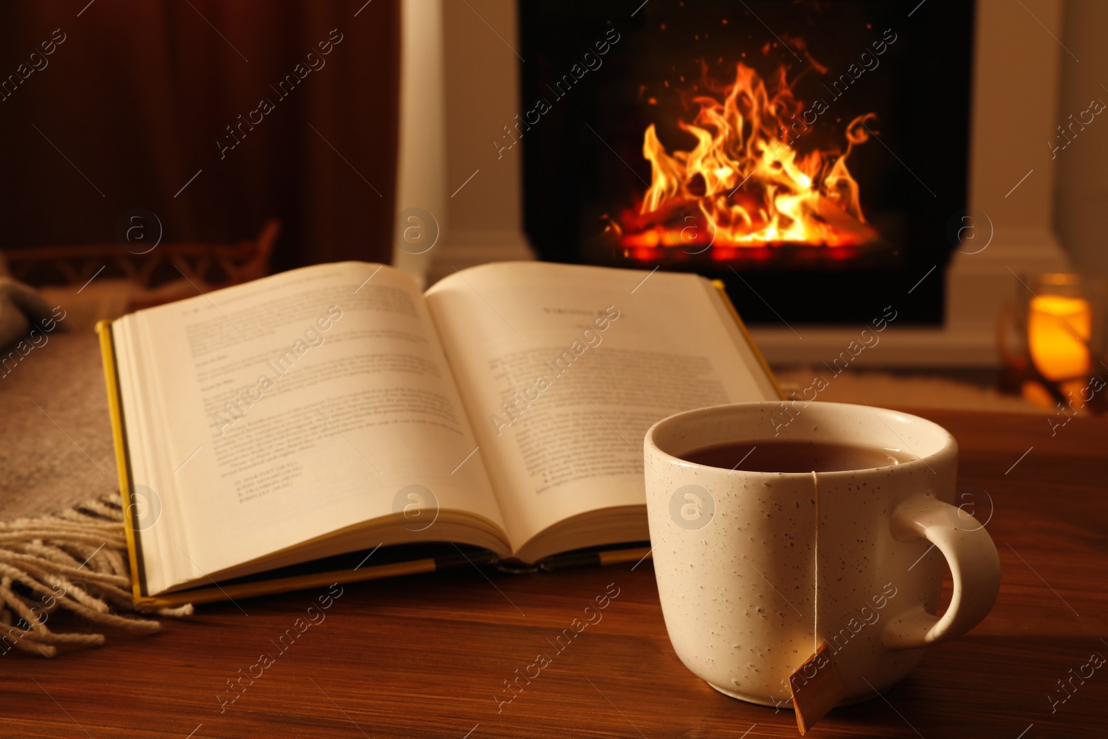 Photo of Cup of hot tea and book on wooden table near fireplace at home. Cozy atmosphere