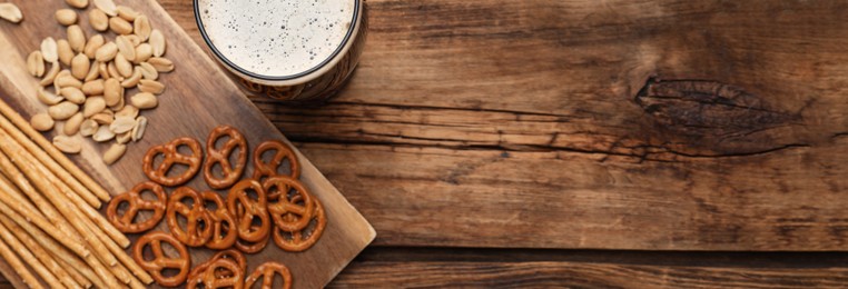 Glass of beer served with delicious pretzel crackers and other snacks on wooden table, flat lay with space for text. Banner design