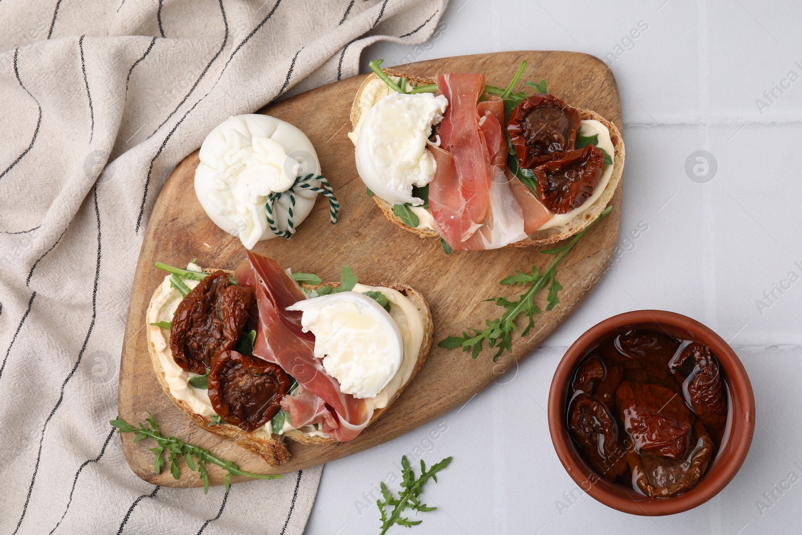 Photo of Delicious sandwiches with burrata cheese, ham and sun-dried tomatoes served on white tiled table, flat lay