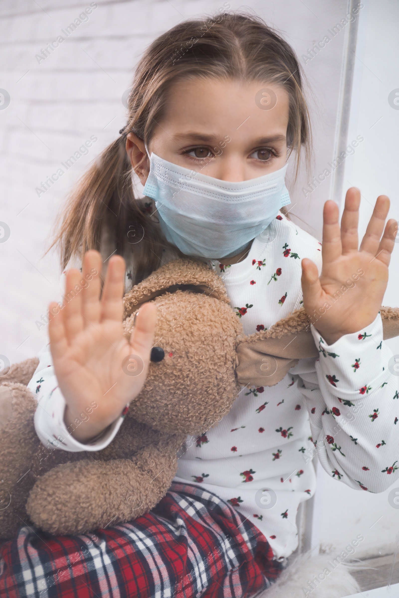 Photo of Sad little girl in protective mask looking out of window indoors, view from outside. Staying at home during coronavirus pandemic