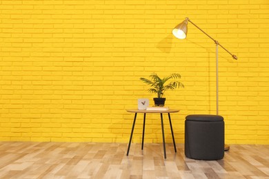 Stylish black pouf, lamp and table near yellow brick wall in room. Space for text