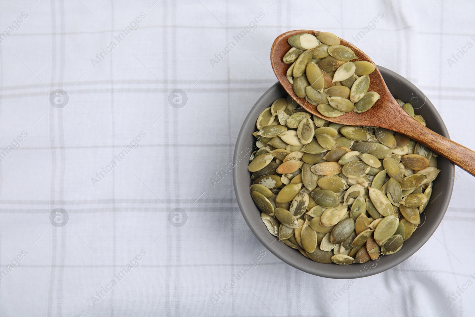 Photo of Bowl with pumpkin seeds and wooden spoon on tablecloth, top view. Space for text