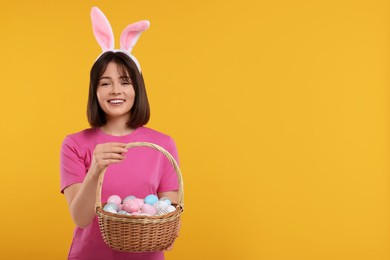 Easter celebration. Happy woman with bunny ears and wicker basket full of painted eggs on orange background, space for text