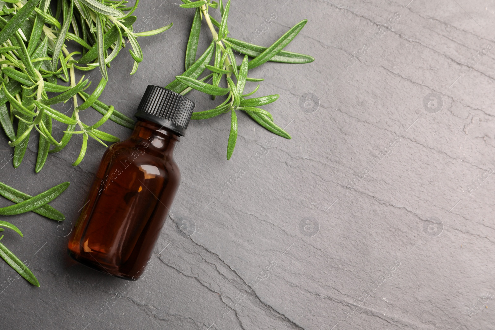 Photo of Bottle of essential oil and fresh rosemary sprigs on gray table, flat lay. Space for text