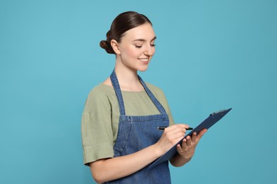 Photo of Beautiful young woman in clean denim apron with clipboard on light blue background