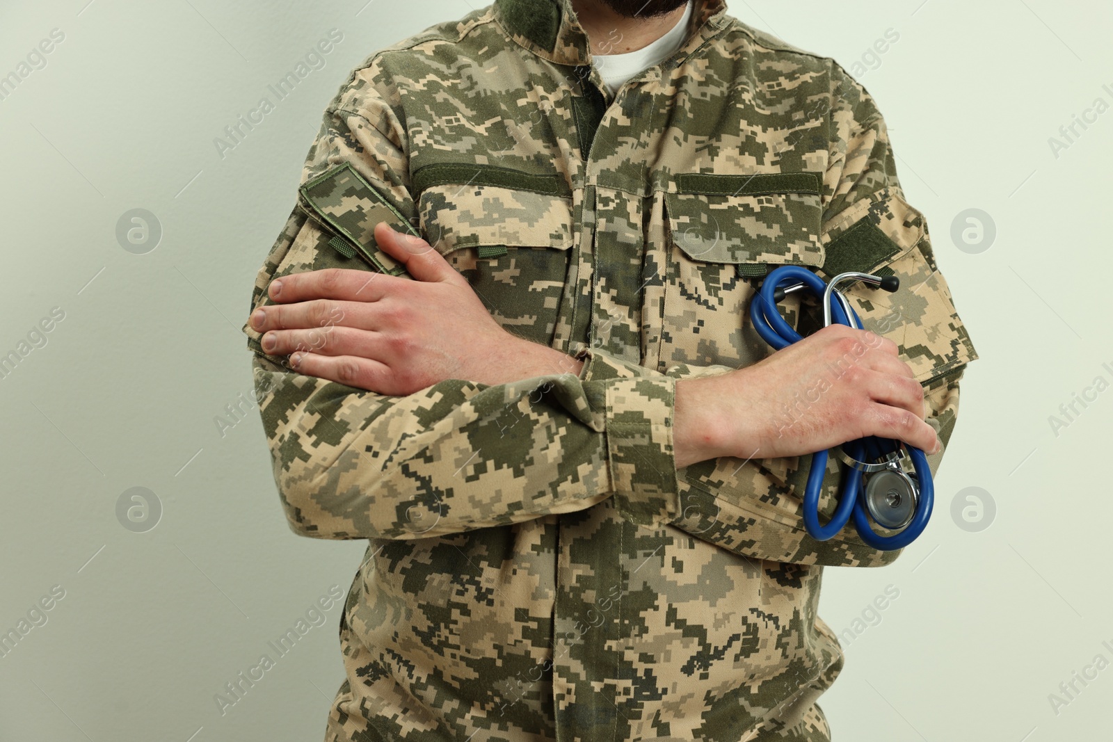 Photo of Man in military uniform with crossed arms and stethoscope on light background, closeup. Health care concept