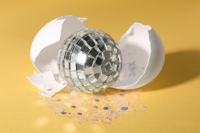 Photo of Silver disco ball, broken eggshell and glitter sprinkles on yellow background, closeup