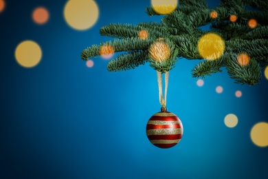 Beautiful Christmas ball hanging on fir tree branch against light blue background, bokeh effect. Space for text