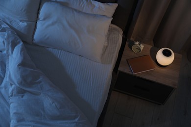 Photo of Nightlight, alarm clock and book on bedside table near bed indoors, above view