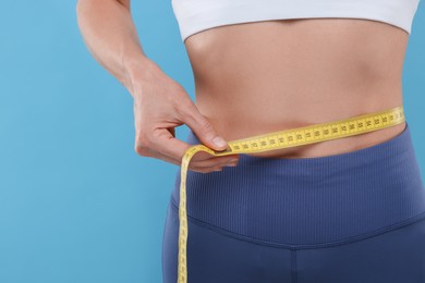 Photo of Slim woman measuring waist with tape on light blue background, closeup. Weight loss