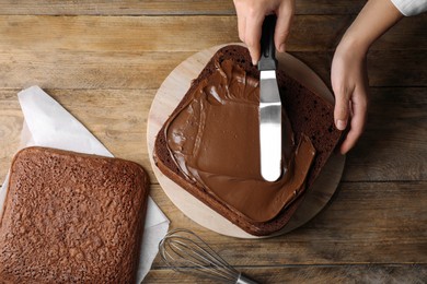 Woman smearing sponge cake with chocolate cream at wooden table, closeup. Top view