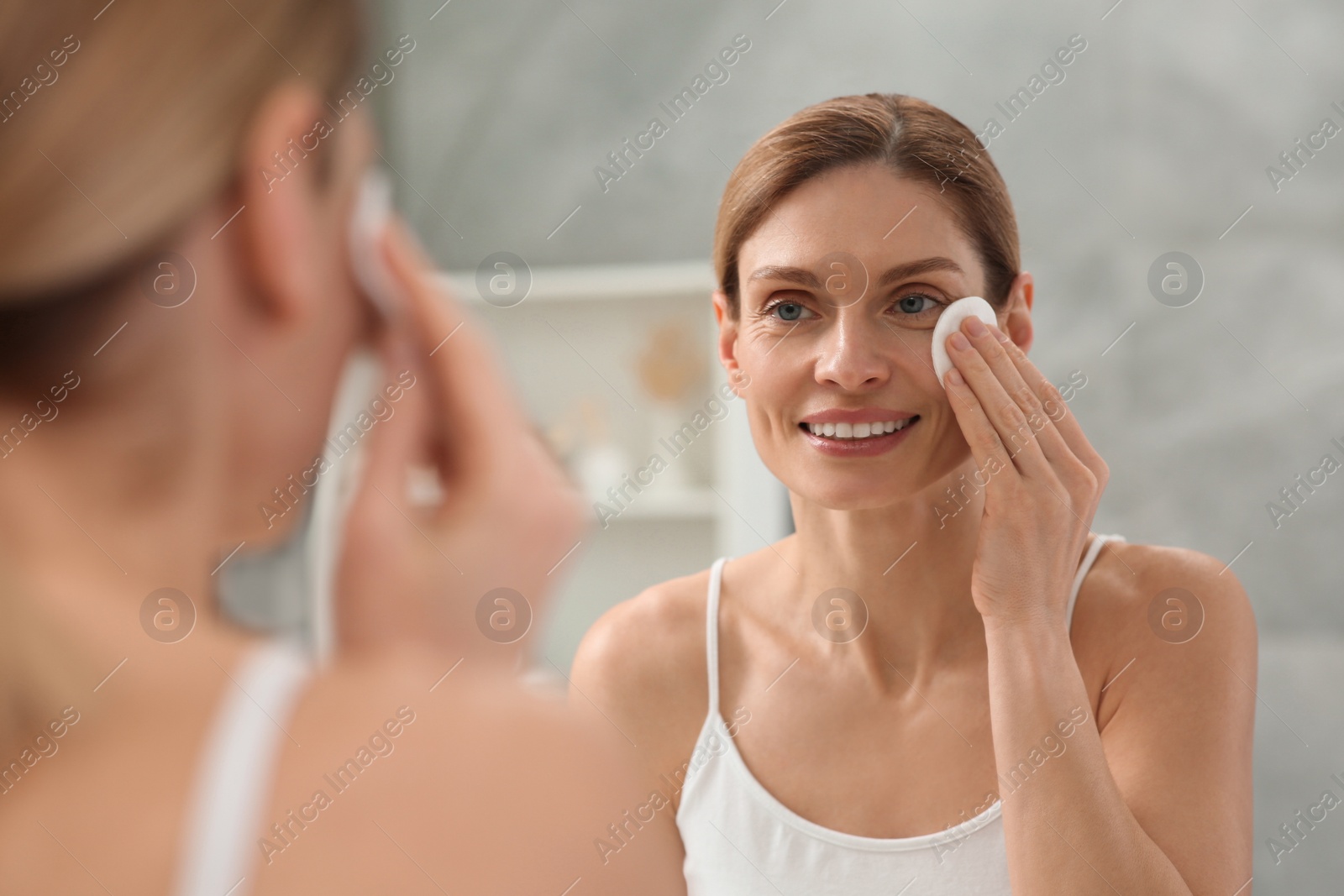 Photo of Beautiful woman removing makeup with cotton pad near mirror indoors