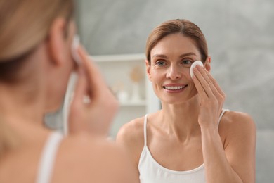 Photo of Beautiful woman removing makeup with cotton pad near mirror indoors