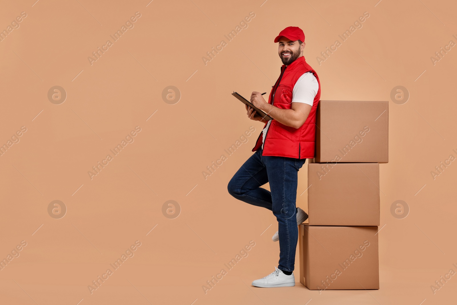 Photo of Happy young courier with clipboard and stack of parcels on light brown background. Space for text