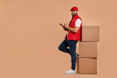Happy young courier with clipboard and stack of parcels on light brown background. Space for text