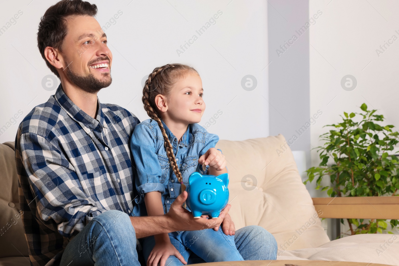 Photo of Little girl with her father putting coin into piggy bank at home, space for text