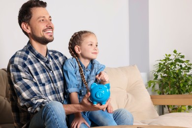 Little girl with her father putting coin into piggy bank at home, space for text