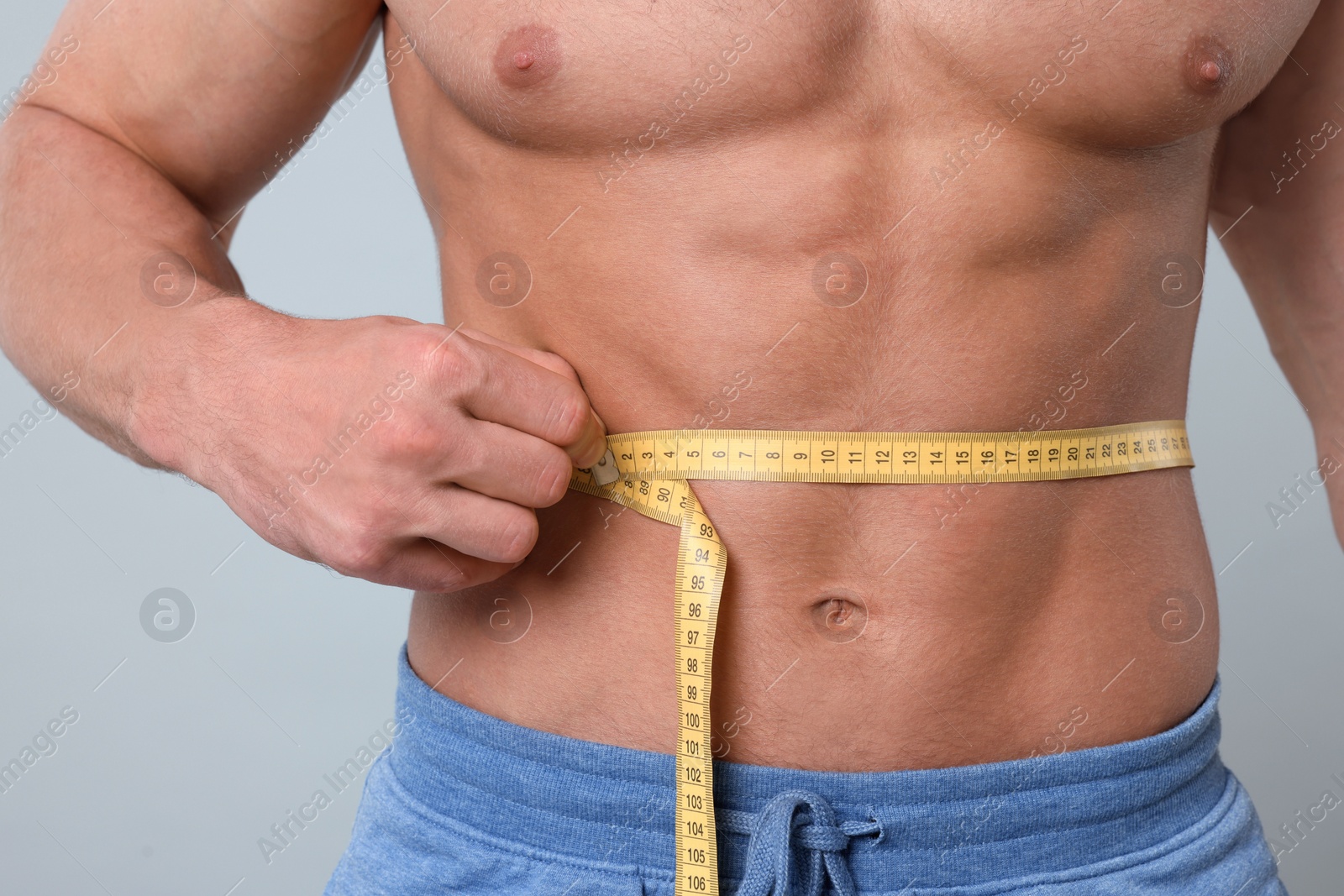 Photo of Athletic man measuring waist with tape on light grey background, closeup. Weight loss concept