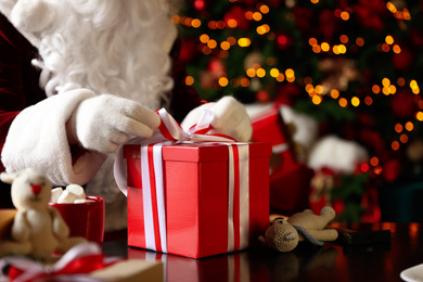 Photo of Santa Claus wrapping Christmas gift against blurred festive lights, closeup