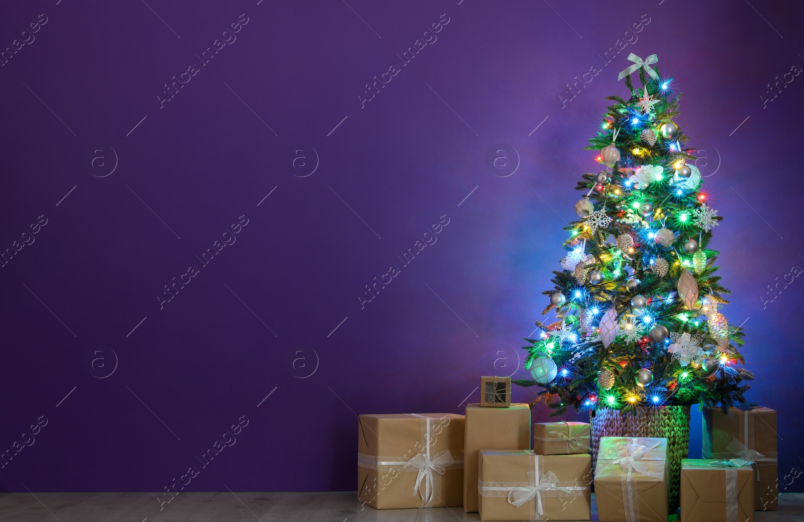 Photo of Beautiful Christmas tree with colorful lights and gift boxes near purple wall indoors, space for text