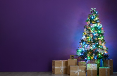 Photo of Beautiful Christmas tree with colorful lights and gift boxes near purple wall indoors, space for text