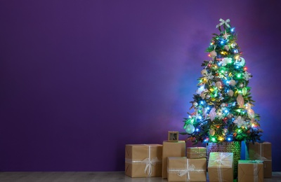 Beautiful Christmas tree with colorful lights and gift boxes near purple wall indoors, space for text