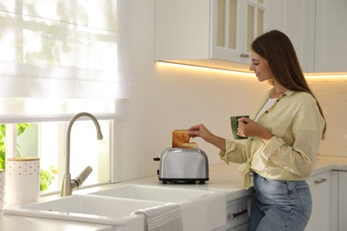 Photo of Young woman taking slice of bread from toaster in kitchen