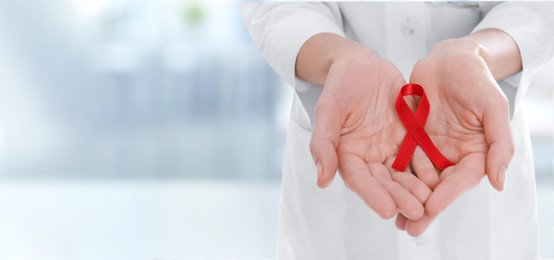 Image of Cancer awareness. Doctor holding red ribbon on blurred background, closeup. Banner design with space for text