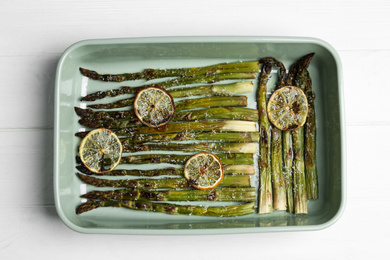 Photo of Oven baked asparagus with lemon slices in ceramic dish on white wooden table, top view