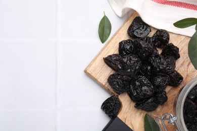 Photo of Tasty dried prunes and green leaves on white tiled table, top view. Space for text