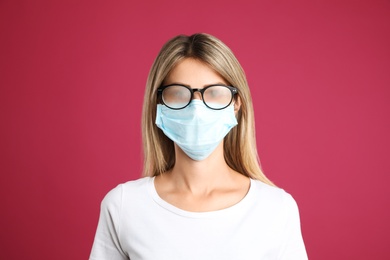 Photo of Woman with foggy glasses caused by wearing disposable mask on pink background. Protective measure during coronavirus pandemic
