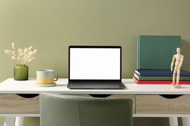 Photo of Stylish workplace with laptop, cup and vase of dry decorative spikes on white desk near olive wall indoors