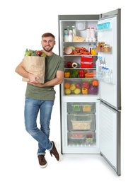 Photo of Young man with bag of groceries near open refrigerator on white background