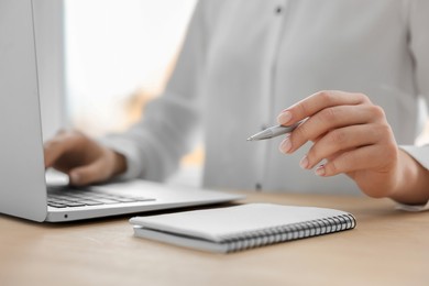 Woman with notebook and pen working on laptop at wooden table, closeup. Electronic document management